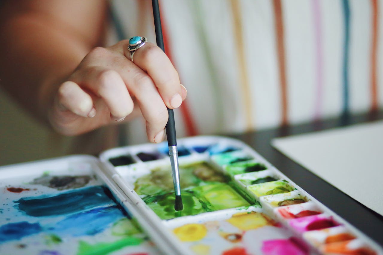 A detailed view of a hand holding a paintbrush, working with vibrant watercolors on a palette.